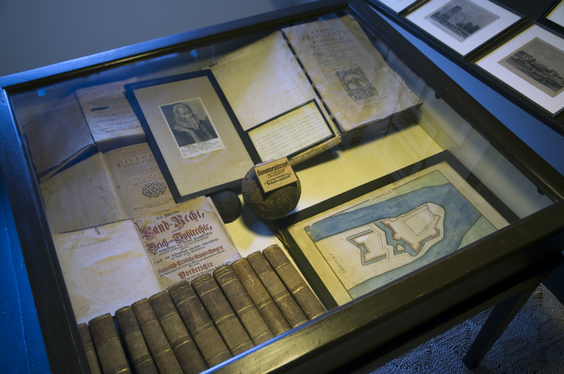 Museum: Vitrine mit historischen Dokumenten und Büchern. Ausgestellt sind Landkarten, Schriftstücke, ein Porträt und weitere Exponate.