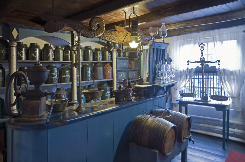 Museum: Historische Ladenansicht. Eine Verkaufstheke mit antiken Waagen, Holzfässern, Teedosen und traditionellen Apothekengläsern, eingerahmt von rustikalen Holzbalken.